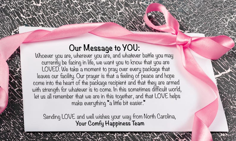 Pu&ograve; includere: Un biglietto bianco con un nastro rosa e il testo "Our Message to YOU:" seguito da un messaggio d'amore e sostegno. Il biglietto &egrave; su una superficie strutturata grigia e nera.