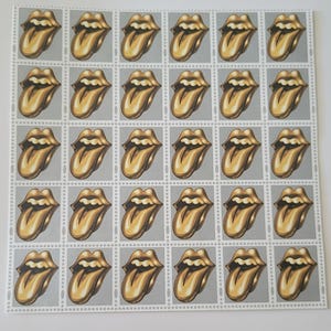 May include: A sheet of 25 gold-coloured postage stamps featuring the iconic Rolling Stones tongue logo. Each stamp is surrounded by a white border and a dotted line.