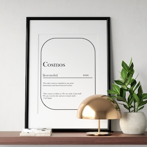 May include: A black framed print with the word "Cosmos" defined as "the entire universe regarded as one great harmonious and interconnected system". The print is on a shelf with a gold lamp and a potted plant.