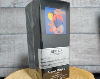 May include: A black bottle of Maison Margiela Replica 'Flying' Eau de Parfum spray. The bottle has a black label with white text and a colorful image of a hot air balloon. The bottle is sitting on a wooden surface.