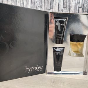 Op de afbeelding: Een zwarte geschenkset met de tekst "hypnôse homme" op de doos. De set bevat twee zwarte tubes "hypnôse" huidverzorgingsproducten en een kleine glazen fles parfum met een zwarte dop.