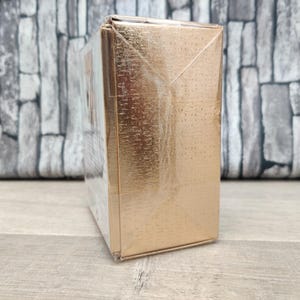 May include: A rectangular, gold-colored box wrapped in clear plastic. The box has a textured surface and is sitting on a wooden surface. The background is a gray brick wall.
