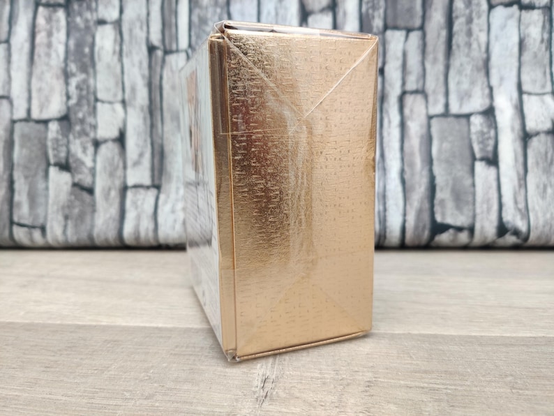 May include: A rectangular, gold-colored box wrapped in clear plastic. The box has a textured surface and is sitting on a wooden surface. The background is a gray brick wall.