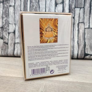 May include: A perfume box with a gold and white design. The front of the box features an illustration of a perfume bottle in a gold and orange color scheme. The box has text in multiple languages.