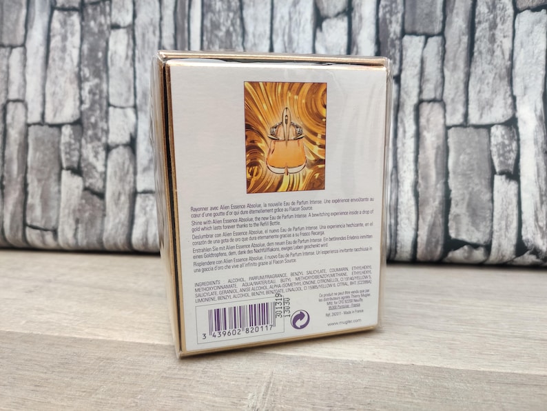 May include: A perfume box with a gold and white design. The front of the box features an illustration of a perfume bottle in a gold and orange color scheme. The box has text in multiple languages.