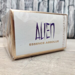 May include: A rectangular box of Alien Essence Absolue perfume. The box is white with gold trim and the word "ALIEN" in large, stylized purple letters. The words "ESSENCE ABSOLUE" are below in gold. The box is wrapped in clear plastic.