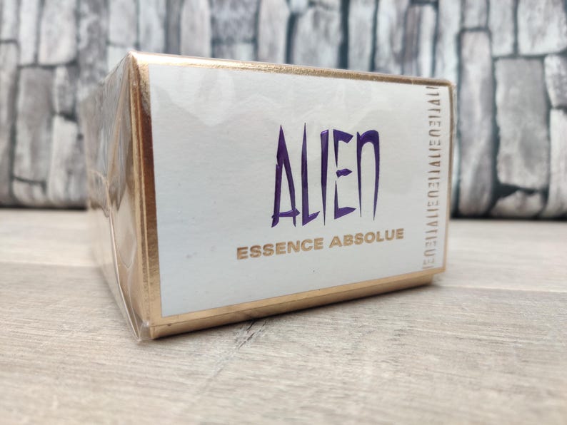 May include: A rectangular box of Alien Essence Absolue perfume. The box is white with gold trim and the word "ALIEN" in large, stylized purple letters. The words "ESSENCE ABSOLUE" are below in gold. The box is wrapped in clear plastic.