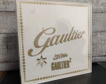 May include: A white box with a gold border and a gold snowflake hanging from the top. The box says "Gaultier" in gold script and "Jean Paul Gaultier" in smaller gold letters. The box also says "Gaultier 2" in gold letters.