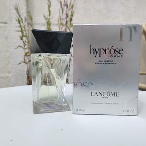 Op de afbeelding: Een heldere glazen parfumfles met een zwarte geometrische dop en een zilveren doos. Op de doos staat "Hypnôse Homme Eau Fraîche Fresh Fragrance" en "Lancôme Paris". De fles bevat 75 ml.