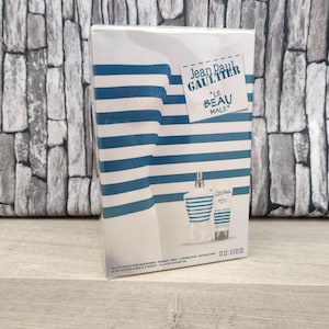 Op de afbeelding: Een doos met Jean Paul Gaultier "Le Beau Male" geur. De verpakking heeft een witte achtergrond met blauwe horizontale strepen. De doos bevat een fles eau de toilette en een bijpassende aftershave balsem.