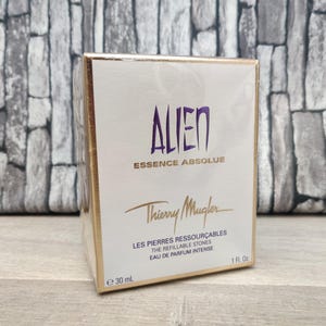 May include: A white and gold box of Thierry Mugler Alien Essence Absolue perfume. The box features the word "ALIEN" in purple, the brand name, and product details. The box contains 1 fl oz (30 ml) of perfume.