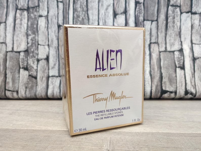 May include: A white and gold box of Thierry Mugler Alien Essence Absolue perfume. The box features the word "ALIEN" in purple, the brand name, and product details. The box contains 1 fl oz (30 ml) of perfume.