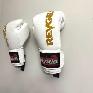 May include: A pair of white boxing gloves with gold REVGEAR branding printed vertically on the exterior. The wrist straps feature a black patch with a red bear logo, the text REVGEAR Original, and a small white tag indicating a size of 12 ounces.