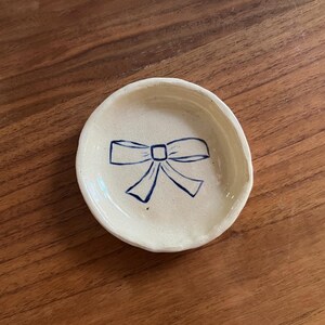 May include: A small, off-white ceramic dish with a hand-drawn blue bow design in the center. The dish has a slightly irregular, organic shape and is set against a warm, wooden surface. Ideal for holding small items.