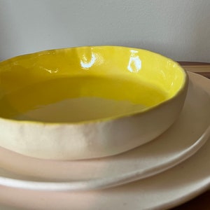 May include: A handmade ceramic bowl with a bright yellow interior and a creamy white exterior, resting on a white plate. The bowl's rim has a slightly irregular shape, adding to its unique, artisanal appeal. The plate below is also white.