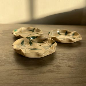 May include: Three small, handmade ceramic dishes with a wavy rim and a beige glaze. Each dish features a hand-painted green leaf design. The dishes are arranged on a wooden surface, with natural light casting shadows.