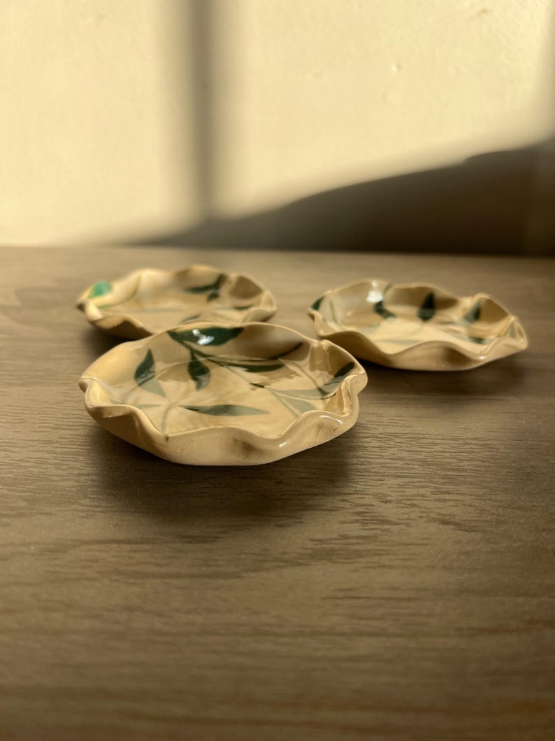 May include: Three small, handmade ceramic dishes with a wavy rim and a beige glaze. Each dish features a hand-painted green leaf design. The dishes are arranged on a wooden surface, with natural light casting shadows.