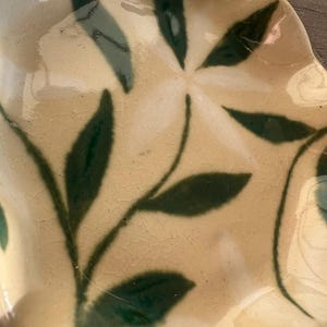 May include: A ceramic plate with a cream-colored base and a dark green leaf design. The plate has a wavy edge and a glossy finish. The leaf pattern is stylized and adds a natural element to the plate's design.