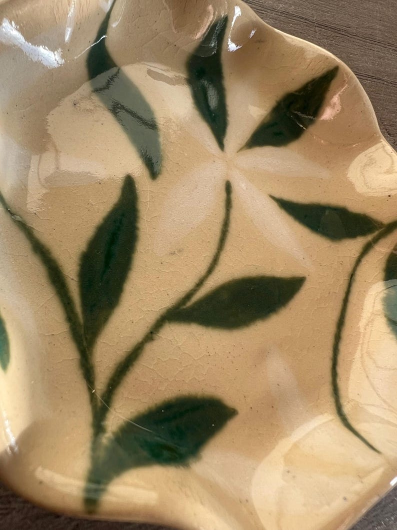 May include: A ceramic plate with a cream-colored base and a dark green leaf design. The plate has a wavy edge and a glossy finish. The leaf pattern is stylized and adds a natural element to the plate's design.