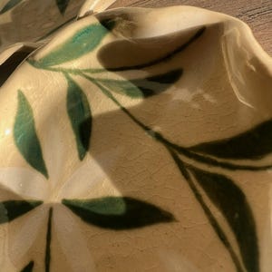 May include: Close-up of two ceramic plates with a cream-colored base and green leaf design. The plates have a wavy edge and a textured surface. The leaves are painted in a dark green color.