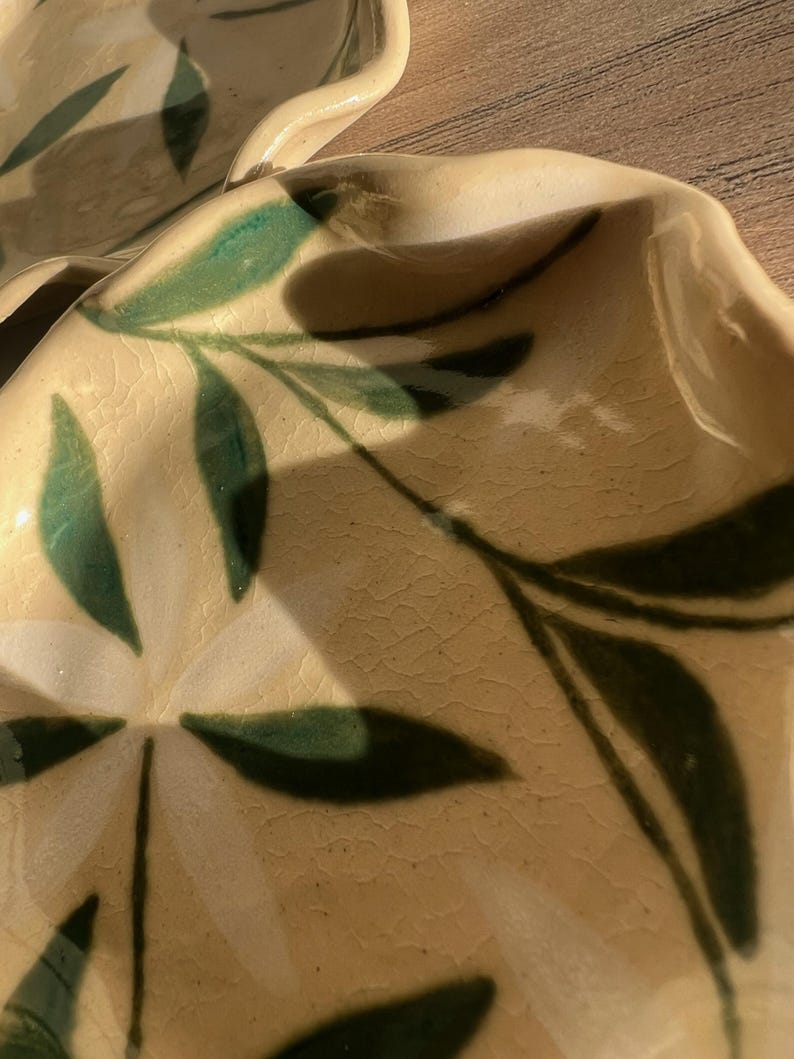 May include: Close-up of two ceramic plates with a cream-colored base and green leaf design. The plates have a wavy edge and a textured surface. The leaves are painted in a dark green color.