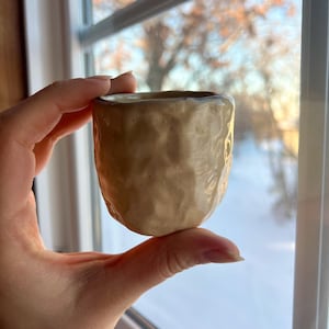 May include: A small, handmade ceramic cup with a textured, beige exterior and a smooth, light brown rim. The cup is held in front of a window, showcasing its unique, organic shape and earthy tones. The cup is approximately 2.5 inches tall.