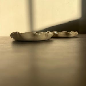 May include: Two small, off-white ceramic dishes with a wavy, organic rim. The dishes are placed on a dark brown surface, with soft lighting and shadows. The dishes appear to be handmade and are likely used for serving small portions or as decorative items.