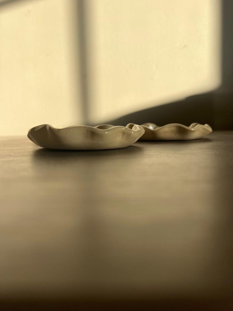 May include: Two small, off-white ceramic dishes with a wavy, organic rim. The dishes are placed on a dark brown surface, with soft lighting and shadows. The dishes appear to be handmade and are likely used for serving small portions or as decorative items.