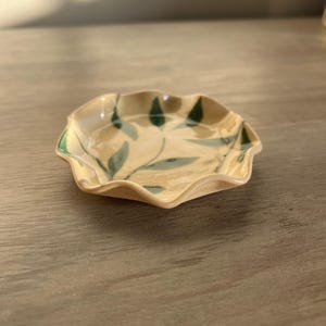 May include: A small, ceramic dish with a scalloped edge. The dish is beige with green leaf designs. The rim is painted green. The dish is likely handmade and suitable for holding small items.