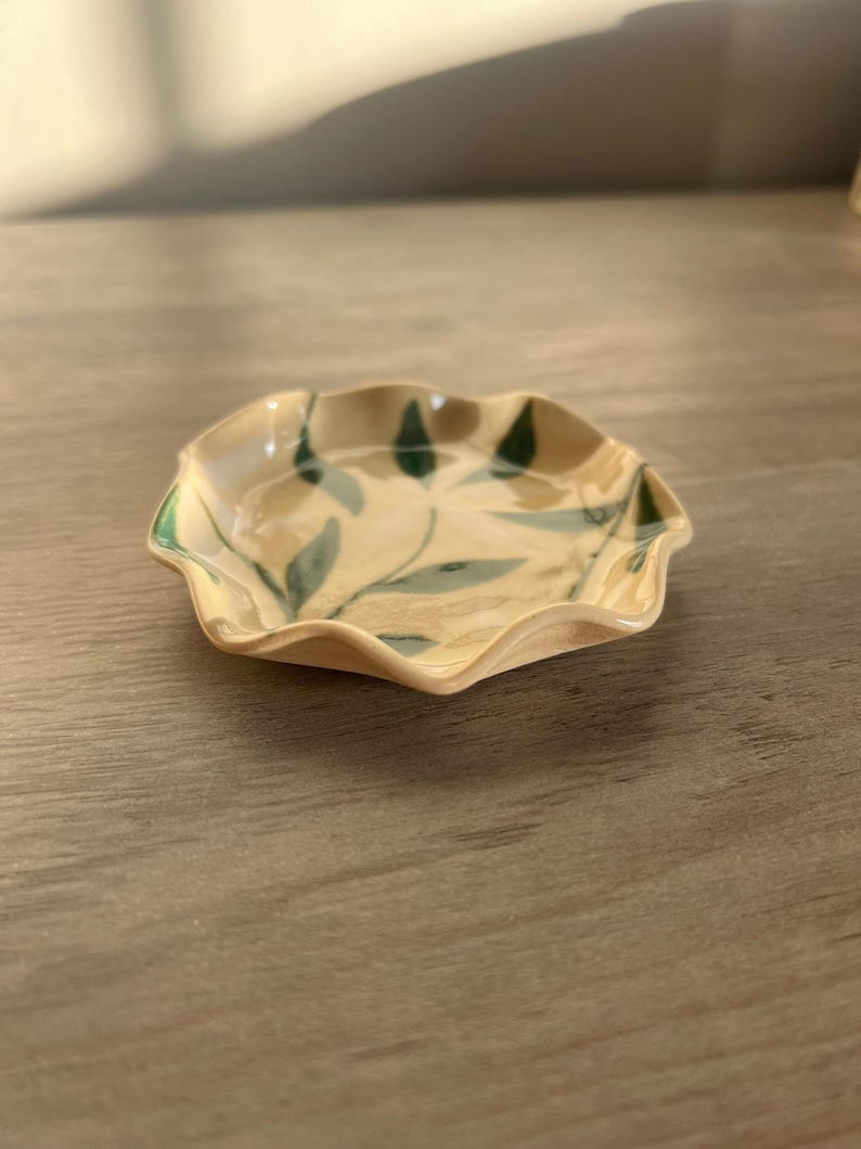 May include: A small, ceramic dish with a scalloped edge. The dish is beige with green leaf designs. The rim is painted green. The dish is likely handmade and suitable for holding small items.