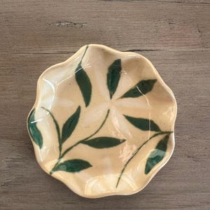 May include: A small, handmade ceramic dish with a scalloped edge. The dish is beige with a green leaf pattern. The leaves are painted in a simple, stylized design. The dish is likely used for serving small portions of food or as a decorative item.