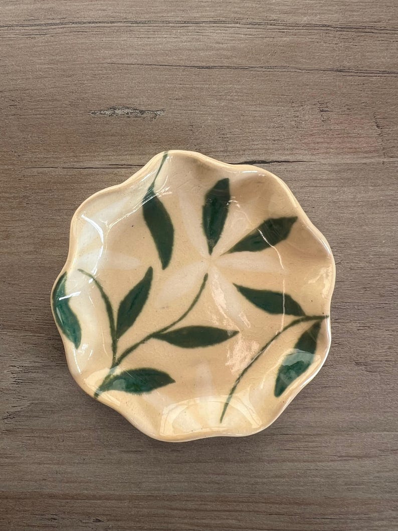 May include: A small, handmade ceramic dish with a scalloped edge. The dish is beige with a green leaf pattern. The leaves are painted in a simple, stylized design. The dish is likely used for serving small portions of food or as a decorative item.