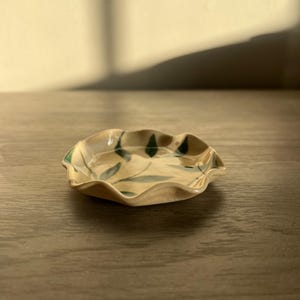 May include: A small, ceramic dish with a ruffled edge. The dish is beige with green leaf designs. The dish is sitting on a wooden surface.