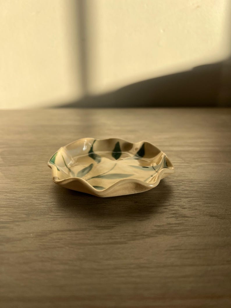 May include: A small, ceramic dish with a ruffled edge. The dish is beige with green leaf designs. The dish is sitting on a wooden surface.