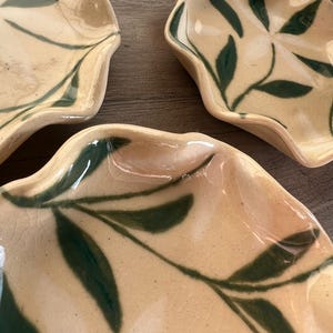 May include: Three ceramic leaf-shaped dishes with a cream-colored base and dark green leaf patterns. The dishes have a glossy finish and are arranged on a wooden surface. The edges of the dishes have a wavy design.