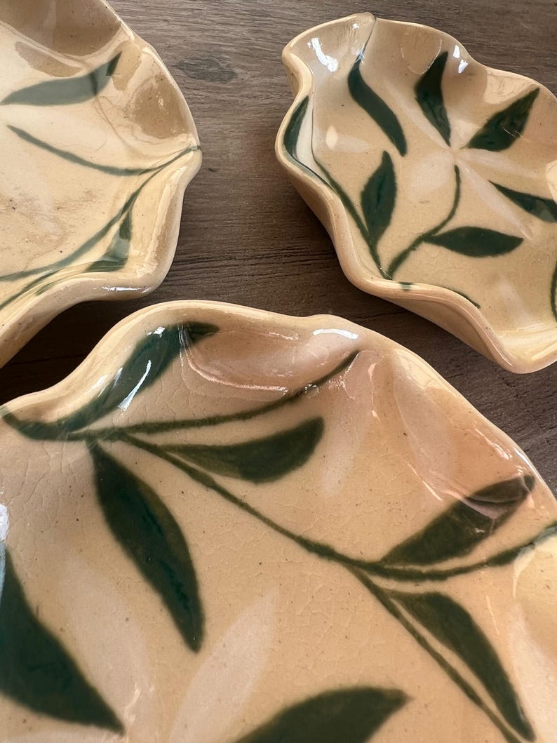 May include: Three ceramic leaf-shaped dishes with a cream-colored base and dark green leaf patterns. The dishes have a glossy finish and are arranged on a wooden surface. The edges of the dishes have a wavy design.