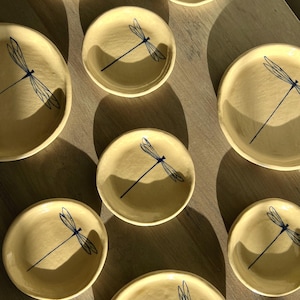 May include: A collection of small, round, cream-colored ceramic dishes. Each dish features a hand-painted blue dragonfly design. The dishes are arranged on a light-colored wooden surface, with shadows adding depth.