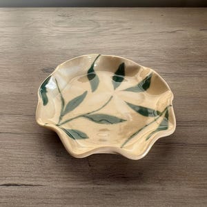 May include: A small, ceramic dish with a scalloped edge. The dish is beige with green leaf designs. The dish is likely used for serving small portions of food or as a decorative item. The dish is on a wooden surface.