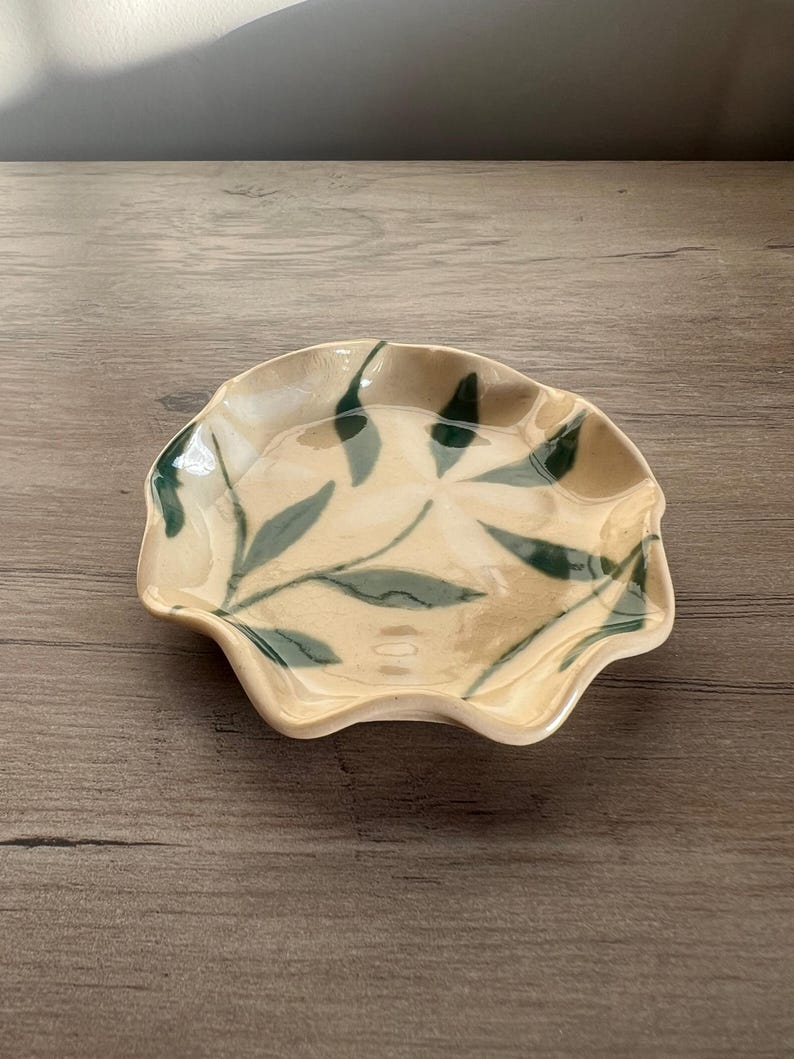 May include: A small, ceramic dish with a scalloped edge. The dish is beige with green leaf designs. The dish is likely used for serving small portions of food or as a decorative item. The dish is on a wooden surface.