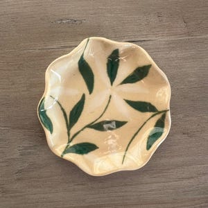 May include: A small, cream-colored ceramic dish with a wavy edge. The dish is decorated with a green leaf pattern. The leaves are arranged in a radial pattern, with each leaf pointing towards the center of the dish. The dish is sitting on a wooden surface.