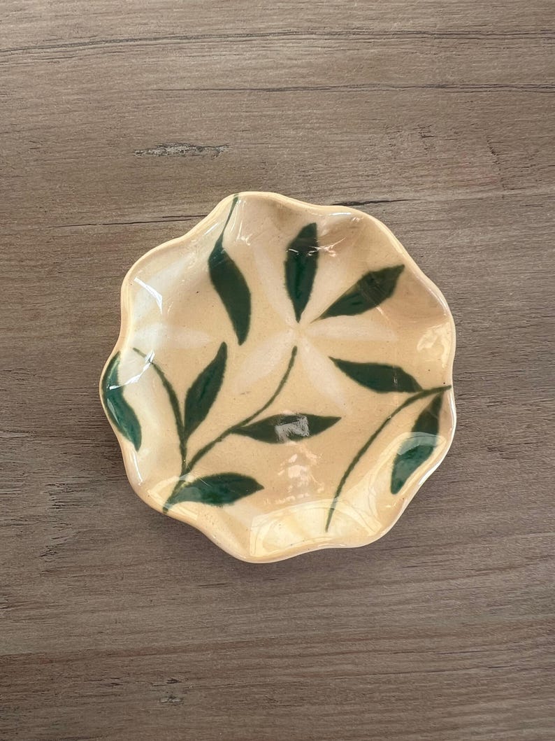 May include: A small, cream-colored ceramic dish with a wavy edge. The dish is decorated with a green leaf pattern. The leaves are arranged in a radial pattern, with each leaf pointing towards the center of the dish. The dish is sitting on a wooden surface.