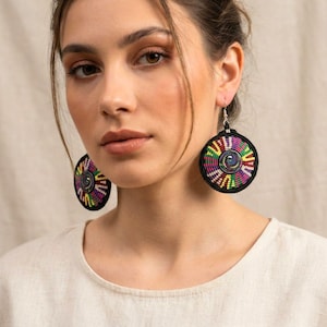 Puede incluir: Pendientes redondos con un diseño tejido en negro, morado, verde, amarillo y rosa. Los pendientes tienen un centro en espiral y están suspendidos de ganchos plateados. Una pieza de joyería llamativa.