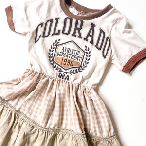 May include: A cream-coloured dress with a brown trim and the word "COLORADO" printed on the front. The dress features a gingham pattern and a tiered skirt. The text "ATHLETIC DEPARTMENT 1990 USA" is also printed on the front.