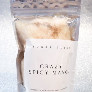 Op de afbeelding: Een zilveren zak met een wit label waarop "Sugar Bliss Crazy Spicy Mango" en "Zuckerwatte mit Mangogesmack & Jalapeno" staat op een witte achtergrond. De zak bevat een pluche, lichtbruine suikerspin.