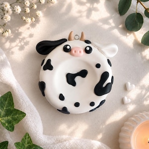 May include: A handmade, white cow-shaped ornament with black spots and accents. The ornament has a pink nose, black eyes, and small horns. It is a decorative item, possibly for a Christmas tree or a similar display.