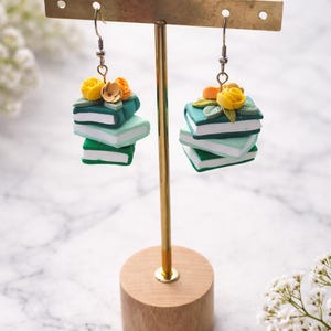 Peut inclure: Boucles d'oreilles représentant des piles de livres miniatures vertes et blanches, ornées de fleurs jaunes et oranges. Les boucles d'oreilles sont présentées sur un support doré avec une base en bois, sur fond de marbre.