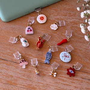 Peut inclure: Collection de breloques de téléphone colorées avec divers motifs : soleil souriant, nuage, cerise, lapin, homard, piment, poisson, champignon, abeille, fleur et gemme. Les breloques sont fixées à des bouchons en plastique transparent.