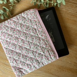 Peut inclure: Une housse de liseuse matelassée à imprimé floral avec une fermeture éclair rose. L'étui est blanc avec un motif de petites fleurs roses et de feuilles vertes. L'e-reader est noir.