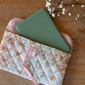 Peut inclure: Une tablette vert sauge avec un logo de flèche incurvée est dans un étui matelassé à motifs floraux. L'étui a une doublure rose et un ruban rose clair. L'étui est blanc avec un motif floral rose, orange et vert. La tablette mesure environ 20 cm de long.