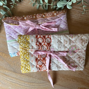 Peut inclure: Deux pochettes en tissu matelassé avec des motifs floraux et géométriques. Les pochettes sont dans les tons de rose, de violet, de jaune et de marron, et sont nouées avec des rubans roses. Elles sont probablement utilisées pour ranger ou transporter de petits objets.